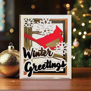 以下が含まれることがあります： 赤いカージナルバードと雪の結晶が描かれた、重ねられた紙のクリスマスカード。「Winter Greetings」の文字が黒い筆記体で書かれています。金色のクリスマスオーナメントが前景にあり、背景にはライトのついたクリスマスツリーがあります。