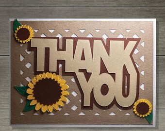 Può includere: Un biglietto d'auguri rettangolare con la scritta THANK YOU in grandi lettere stampatello color marrone chiaro con un'ombra marrone scuro. Lo sfondo presenta un motivo a griglia marrone chiaro, decorato con tre girasoli dai petali gialli.