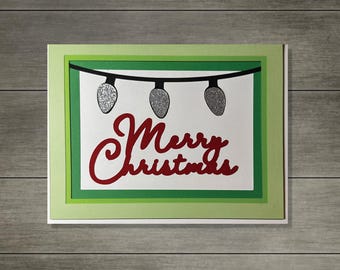 May include: A handmade Christmas card with a layered design. The card features a white background with a green border, and a red script that says "Merry Christmas". Three silver glitter lights hang from a black string.