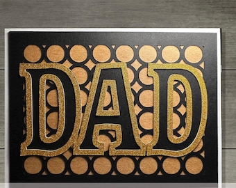 May include: A rectangular greeting card featuring the word DAD in large, bold, gold glitter letters with black outlines. The background consists of a black die-cut layer with a repeating circular pattern revealing a tan, textured paper underneath.