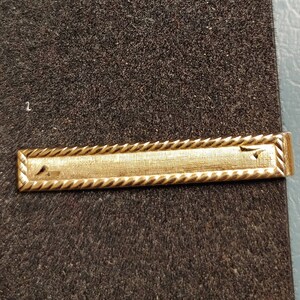 May include: A gold-colored tie bar with a rope-like design around the edges.