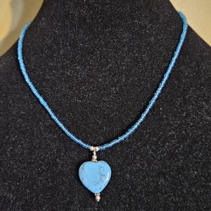 May include: A blue beaded necklace with a heart-shaped pendant. The pendant is a light blue stone with a silver bail. The necklace is displayed on a black velvet bust.