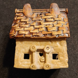 May include: A small ceramic house with a brown roof and a white body. The house has a heart-shaped door and two windows.