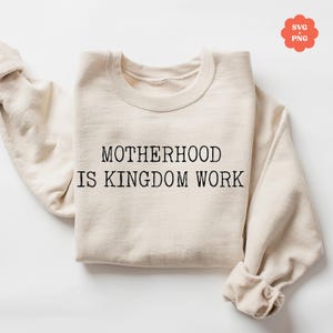 May include: A beige sweatshirt with the text "MOTHERHOOD IS KINGDOM WORK" printed in black.