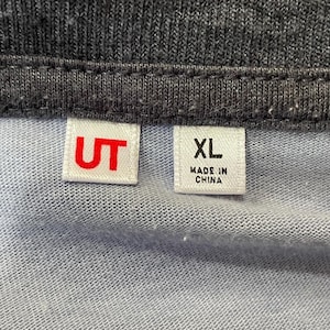 Może przedstawiać: Zbliżenie jasnoniebieskiej i szarej tkaniny z dwiema białymi etykietami. Jedna etykieta ma czerwone litery "UT", a druga etykieta ma "XL" i "MADE IN CHINA" wydrukowane na czarno.