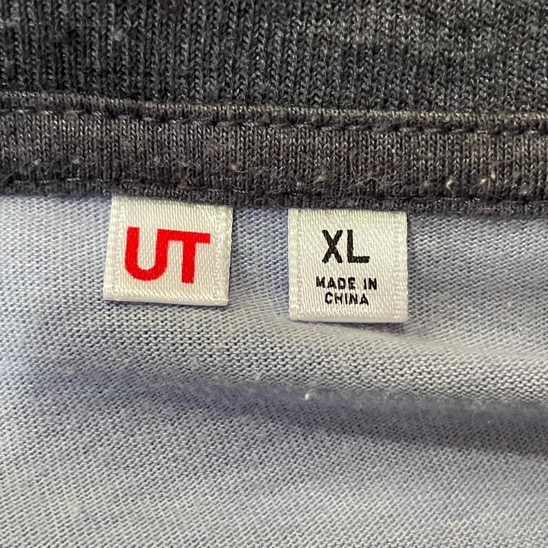 Może przedstawiać: Zbliżenie jasnoniebieskiej i szarej tkaniny z dwiema białymi etykietami. Jedna etykieta ma czerwone litery "UT", a druga etykieta ma "XL" i "MADE IN CHINA" wydrukowane na czarno.