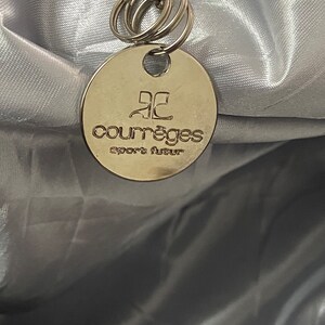 May include: A silver round tag with the text "Courr&egrave;ges Sport Fluer" on it. The tag is attached to a silver chain with multiple silver rings.