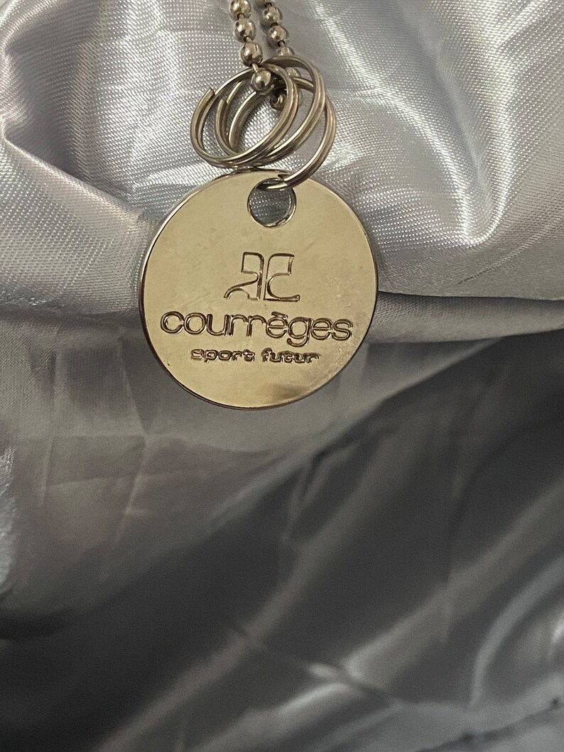 May include: A silver round tag with the text "Courr&egrave;ges Sport Fluer" on it. The tag is attached to a silver chain with multiple silver rings.