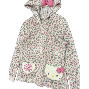 May include: A pink and white fluffy zip-up hoodie with a leopard print. The hoodie has a pink bow on the hood and a Hello Kitty patch on the front.