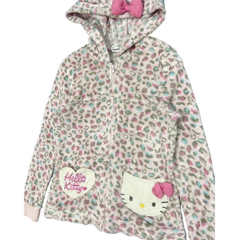 May include: A pink and white fluffy zip-up hoodie with a leopard print. The hoodie has a pink bow on the hood and a Hello Kitty patch on the front.