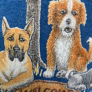 May include: A blue sweatshirt with a graphic of two dogs and a sign that says "Welcome". The dogs are brown and white with black noses. The sign is brown with white lettering.