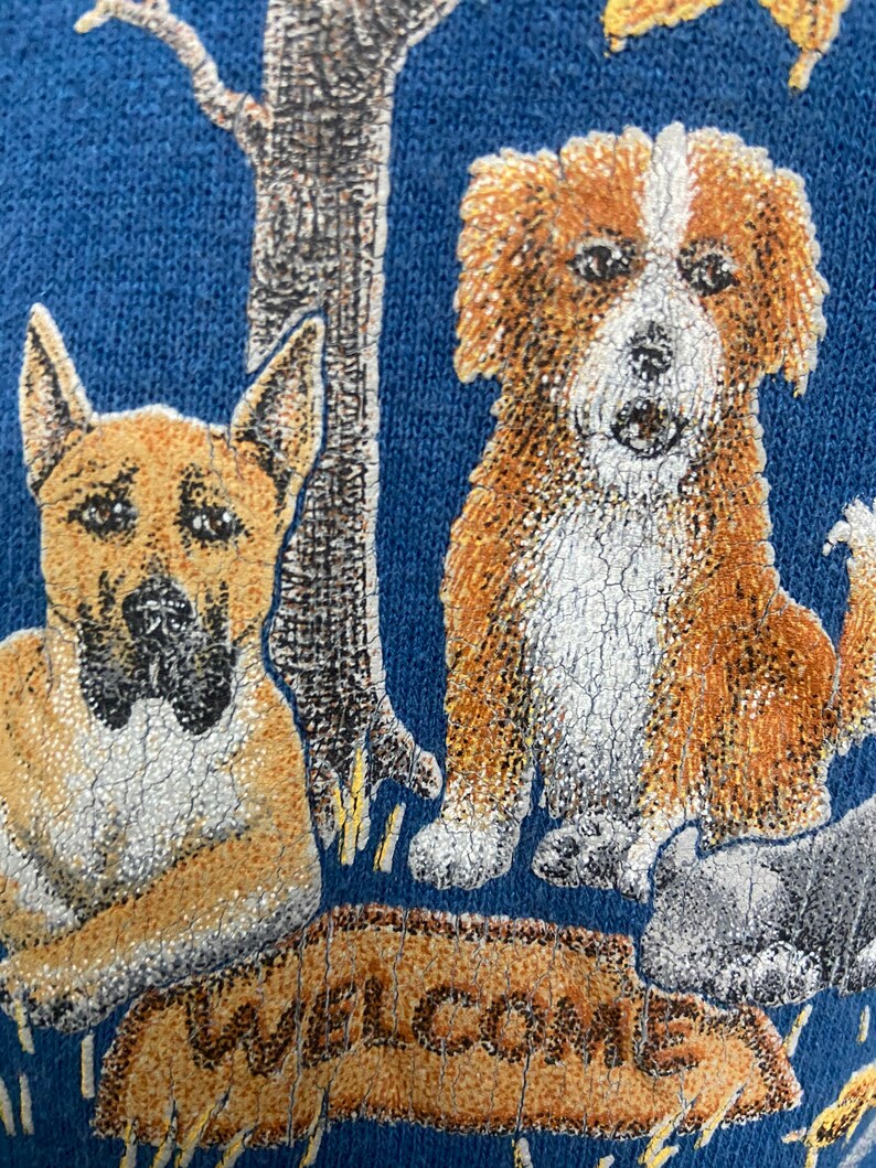 May include: A blue sweatshirt with a graphic of two dogs and a sign that says "Welcome". The dogs are brown and white with black noses. The sign is brown with white lettering.