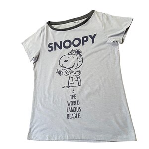 Może przedstawiać: Jasnoszara koszulka z granatowym napisem "SNOOPY" i kresk&oacute;wkową ilustracją Snoopy'ego w czapce pilota. Tekst "IS THE WORLD FAMOUS BEAGLE" znajduje się poniżej. Koszulka ma ciemnoszare wykończenie.