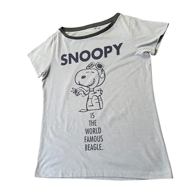 Może przedstawiać: Jasnoszara koszulka z granatowym napisem "SNOOPY" i kresk&oacute;wkową ilustracją Snoopy'ego w czapce pilota. Tekst "IS THE WORLD FAMOUS BEAGLE" znajduje się poniżej. Koszulka ma ciemnoszare wykończenie.
