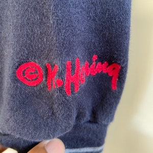 Może przedstawiać: Koszulka z długim rękawem w kolorze granatowym z haftowanym czerwonym logo z napisem "K-Swiss" i czerwonym k&oacute;łkiem z ozdobnym "e" w środku.