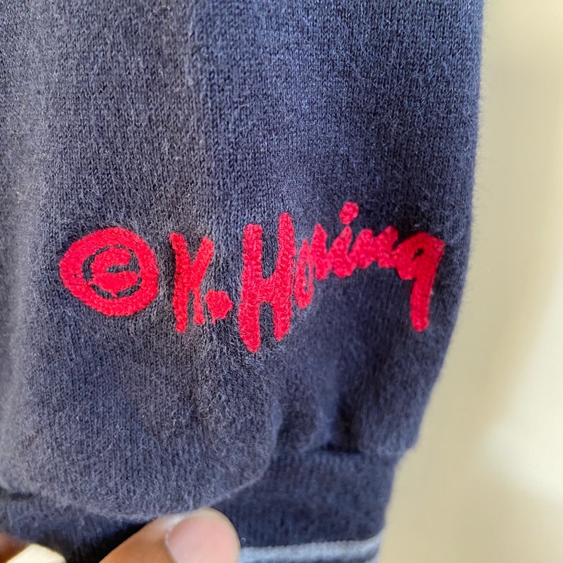 Może przedstawiać: Koszulka z długim rękawem w kolorze granatowym z haftowanym czerwonym logo z napisem "K-Swiss" i czerwonym k&oacute;łkiem z ozdobnym "e" w środku.