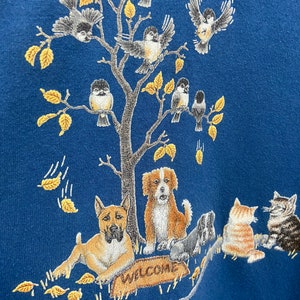 May include: A blue sweatshirt with an embroidered design of a tree with birds, a dog, and a cat. The tree has yellow leaves and the animals are in various poses. The text "WELCOME" is embroidered on a sign at the base of the tree.