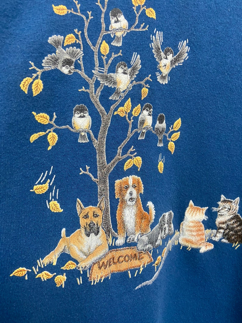 May include: A blue sweatshirt with an embroidered design of a tree with birds, a dog, and a cat. The tree has yellow leaves and the animals are in various poses. The text "WELCOME" is embroidered on a sign at the base of the tree.