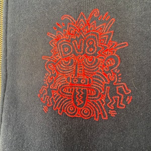 Może przedstawiać: Czerwony projekt graficzny na czarnym tle. Projekt przedstawia stylizowaną twarz z napisem "DV8" w środku.