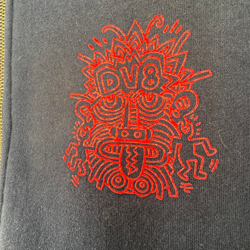 Może przedstawiać: Czerwony projekt graficzny na czarnym tle. Projekt przedstawia stylizowaną twarz z napisem "DV8" w środku.