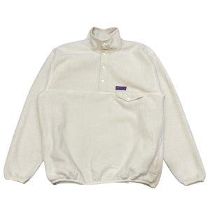May include: Cream-coloured fleece pullover with a high collar and button closure. It has a small chest pocket and a rectangular patch with text. The long-sleeved top is made of a soft, textured material.