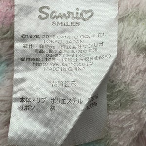 May include: A white tag with black text, including the Sanrio logo and the text "&copy;1976, 2013 SANRIO CO., LTD. TOKYO, JAPAN". The tag also includes information about the product's composition, including "Polyester 100%" and "Cotton 100%".