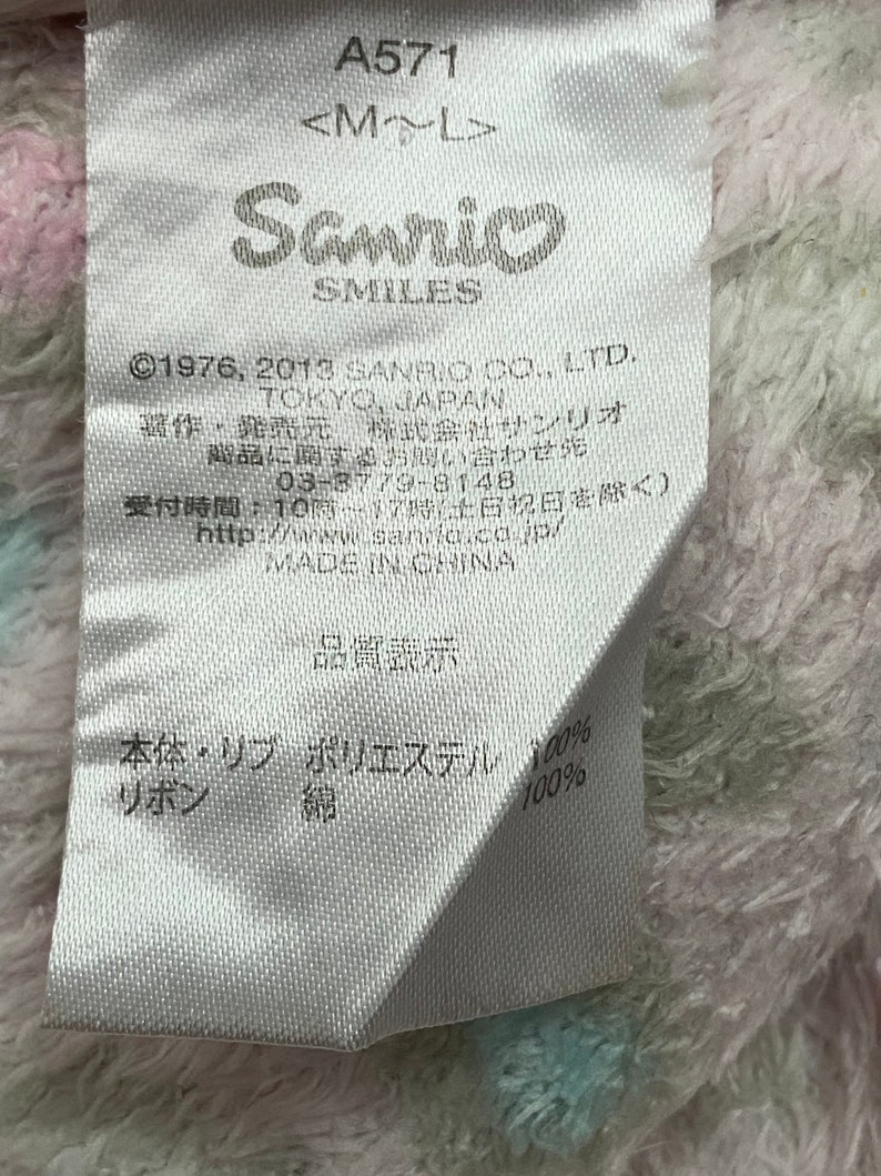 May include: A white tag with black text, including the Sanrio logo and the text "&copy;1976, 2013 SANRIO CO., LTD. TOKYO, JAPAN". The tag also includes information about the product's composition, including "Polyester 100%" and "Cotton 100%".