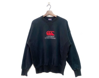 Puede incluir: Sudadera negra con un logotipo rojo y blanco de Canterbury de Nueva Zelanda.