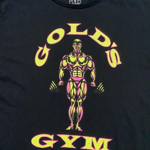 Op de afbeelding: Zwart T-shirt met een gele en roze afbeelding van een bodybuilder die gewichten heft. De afbeelding zegt "Gold's Gym".