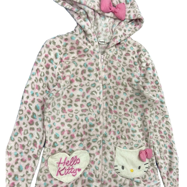 May include: A pink and white fleece zip-up hoodie with a leopard print. The hoodie has a hood with a pink bow, a Hello Kitty heart patch on the front, and a Hello Kitty pocket on the front.