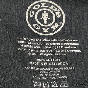 Op de afbeelding: Een zwart label met witte tekst die "Large", "Gold's Gym", "100% Katoen", "Gemaakt in El Salvador" en wasinstructies aangeeft. Het label bevat ook het Gold's Gym-logo, dat een cirkel is met een gewichtheffer erin.