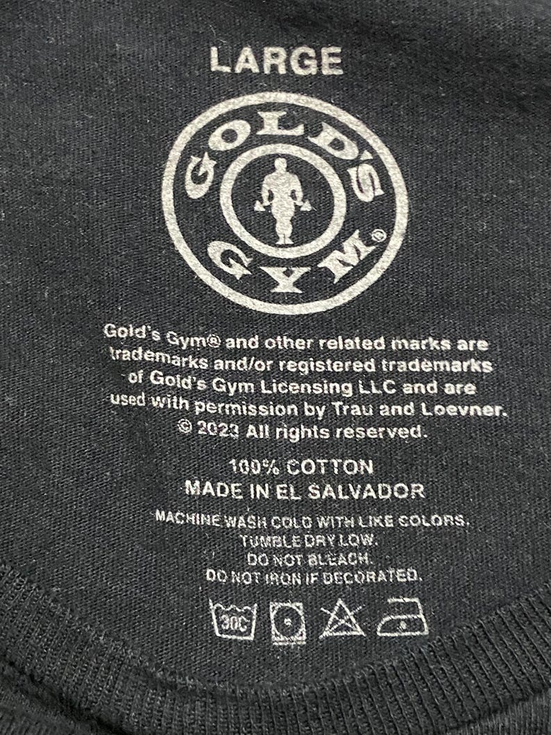 Op de afbeelding: Een zwart label met witte tekst die "Large", "Gold's Gym", "100% Katoen", "Gemaakt in El Salvador" en wasinstructies aangeeft. Het label bevat ook het Gold's Gym-logo, dat een cirkel is met een gewichtheffer erin.
