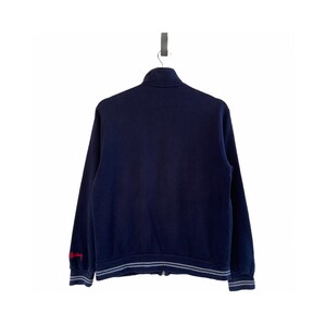 Może przedstawiać: Granatowa bluza zapinana na zamek błyskawiczny z białymi prążkowanymi wykończeniami wok&oacute;ł mankiet&oacute;w i pasa. Bluza ma haftowany czerwony napis "Off-White" na lewym rękawie.