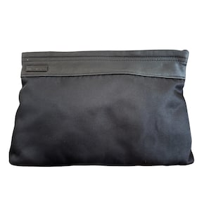 Op de afbeelding: Een rechthoekige zwarte nylon clutch met een gladde donkerbruine lederen bies langs de bovenrand. De tas heeft een minimalistisch ontwerp met een klein rechthoekig lederen lipje aan de linkerbovenkant, wat zorgt voor een strakke en functionele uitstraling.