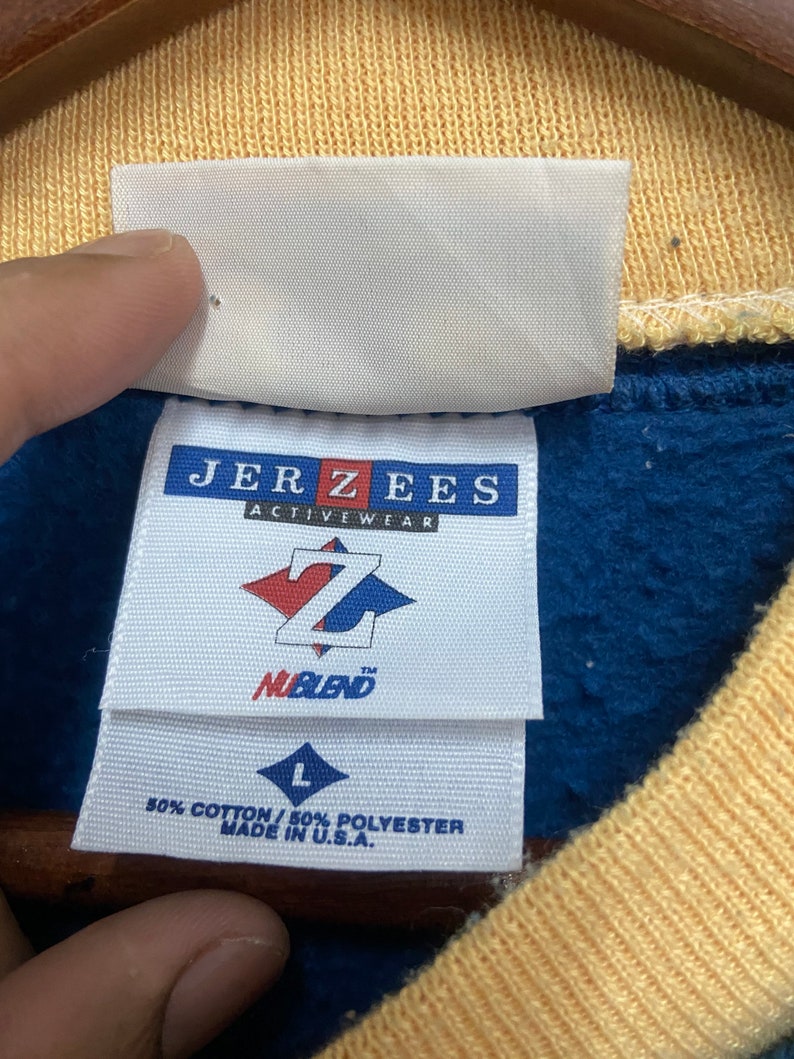 May include: A blue and yellow Jerzees sweatshirt tag with the text "Jerzees Activewear" and a Z logo with the text "Nablend". The tag also includes the size "L" and the text "80% Cotton / 50% Polyester Made in U.S.A."
