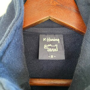 Może przedstawiać: Granatowa bluza z kapturem z czarną metką z napisem "K. Haring" i białym nadrukiem psa. Metka zawiera r&oacute;wnież napis "S."