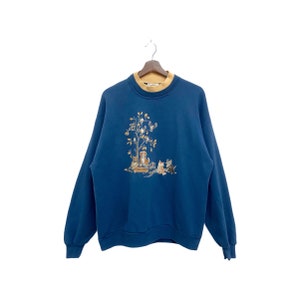 May include: A blue sweatshirt with a brown and white embroidered design of a tree with birds and animals. The text "Welcome" is on a sign at the base of the tree.