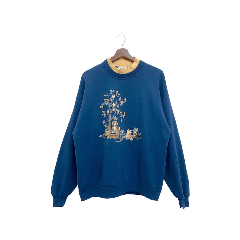 May include: A blue sweatshirt with a brown and white embroidered design of a tree with birds and animals. The text "Welcome" is on a sign at the base of the tree.