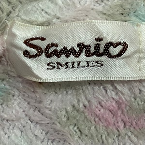 May include: White fabric with a pink and green stripe pattern. A white label with a brown "Sanrio Smiles" logo is attached to the fabric.