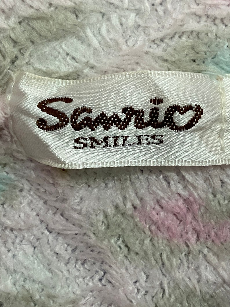 May include: White fabric with a pink and green stripe pattern. A white label with a brown "Sanrio Smiles" logo is attached to the fabric.