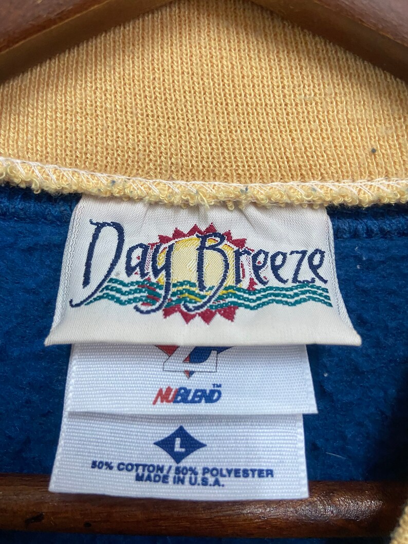 May include: A blue and yellow tag with the text "Day Breeze" and a sun graphic. Below the tag is a white tag with the text "NuBlend" and a size "L" tag. The tag also says "50% Cotton / 50% Polyester Made in U.S.A."