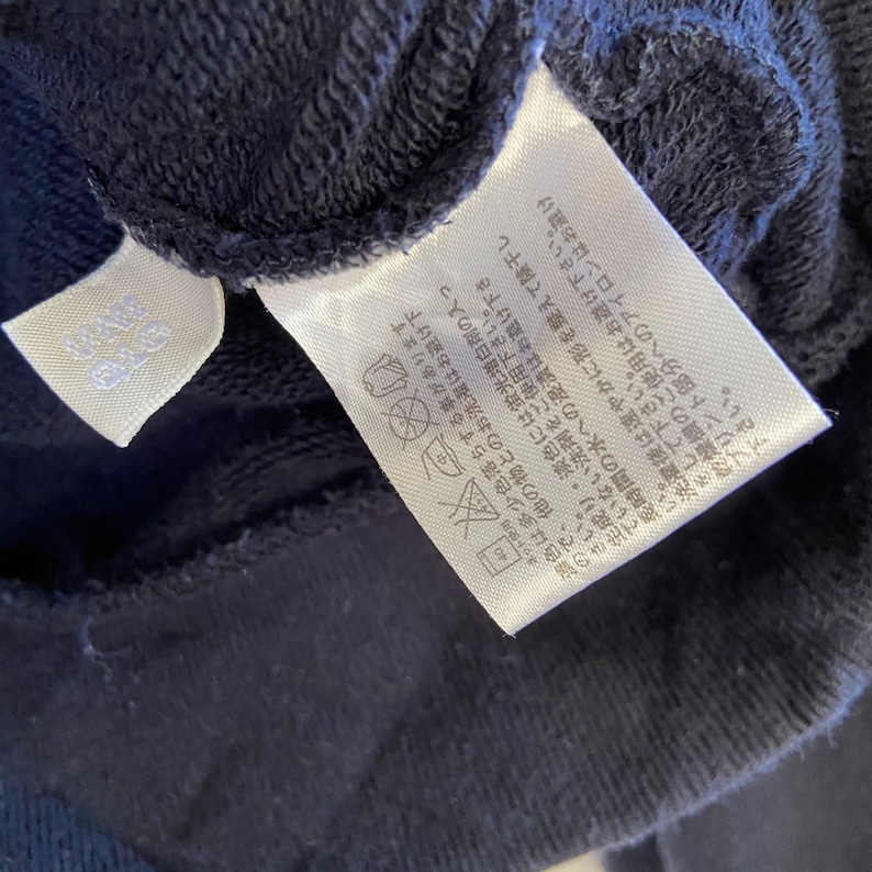 Może przedstawiać: Zbliżenie na ciemnoniebieską metkę pielęgnacyjną z japońskim tekstem i instrukcjami prania. Metka zawiera symbole prania w pralce, nie wybielać, nie suszyć w suszarce bębnowej i prasować w niskiej temperaturze.