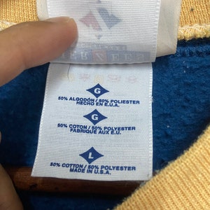 May include: A white tag with black text indicating the size and fabric content of a garment. The tag reads "G 50% ALGOD&Oacute;N / 50% POLIESTER HECHO EN E.U.A. G 50% COTON/50% POLYESTER FABRIQUE AUX E.U. L 50% COTTON/50% POLYESTER MADE IN U.S.A."