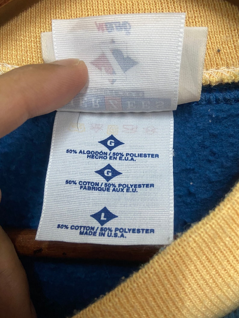 May include: A white tag with black text indicating the size and fabric content of a garment. The tag reads "G 50% ALGOD&Oacute;N / 50% POLIESTER HECHO EN E.U.A. G 50% COTON/50% POLYESTER FABRIQUE AUX E.U. L 50% COTTON/50% POLYESTER MADE IN U.S.A."