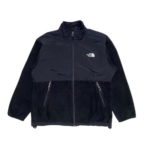 May include: A black fleece jacket with a zipper closure and a black nylon panel on the chest. The jacket has a white logo that reads "The North Face" on the chest.