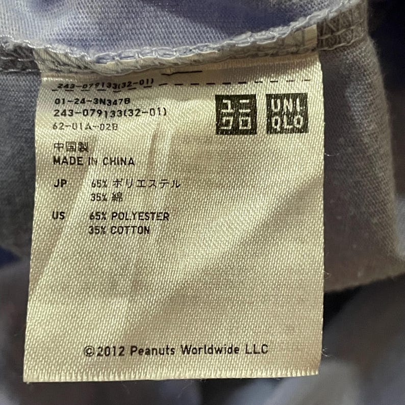 Może przedstawiać: Zbliżenie metki odzieżowej z logo UNIQLO i tekstem. Etykieta jest beżowa z czarnym tekstem wskazującym skład: 65% poliester, 35% bawełna. Etykieta zawiera r&oacute;wnież tekst "MADE IN CHINA" i "&copy;2012 Peanuts Worldwide LLC."