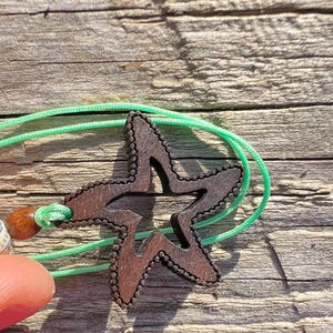 Könnte beinhalten: Ein Holz-Seestern-Anhänger mit einem Ausschnittmuster, der an einer grünen Schnur hängt.