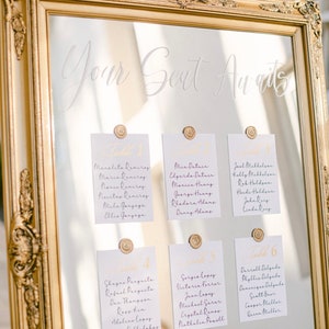 May include: A gold-framed mirror with a white inscription "Your Seat Awaits" and cards with names and table numbers attached with gold wax seals.