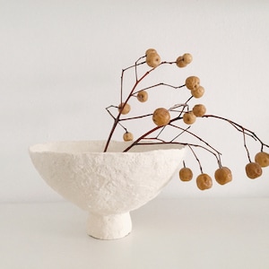 Puede incluir: Un cuenco de cerámica blanca con una superficie texturizada y una base ancha y redondeada. El cuenco está lleno de una rama de bayas secas.