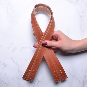 May include: Two brown leather straps with stitching detail, held in a hand. The straps are folded in half and crossed over each other.